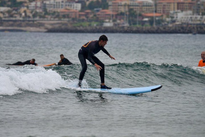 Een surfer die een golf pakt, met andere surfers op de achtergrond en een kuststreeed. 2