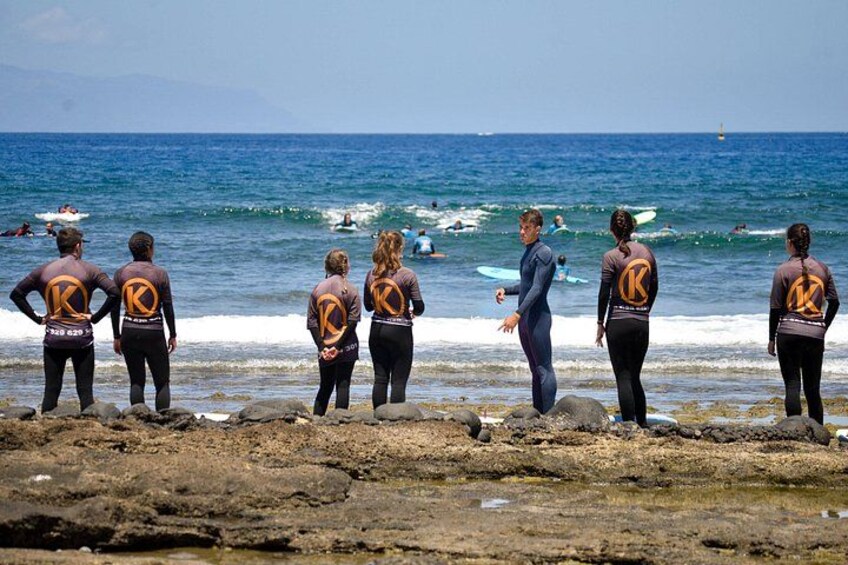 Een groep mensen in neopreenpakken staat op rotsen aan de oceaan en kijkt naar surfers. 5