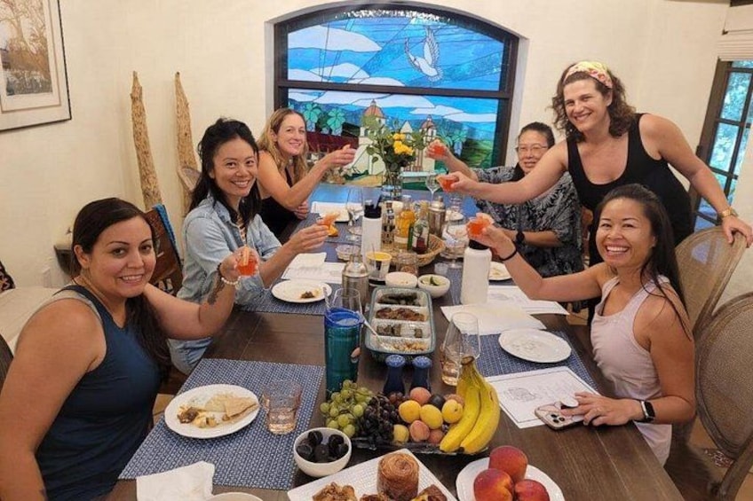 Un grupo de personas sentadas alrededor de una mesa con comida y bebidas, una ventana con vistas y un cuadro en la pared. 3