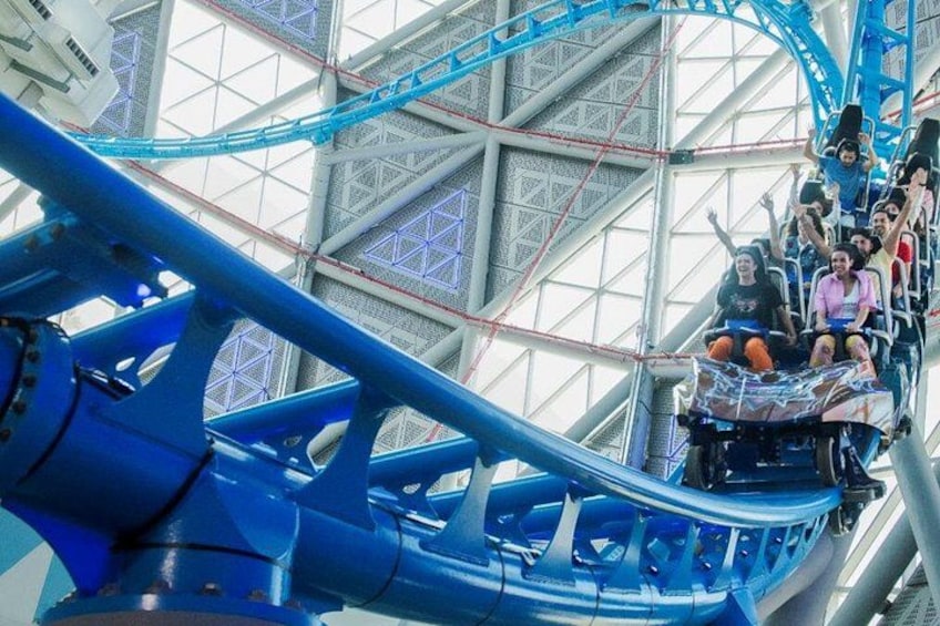 Una montaña rusa dentro de un parque de atracciones techado, con estructura de acero y rieles azules. 1