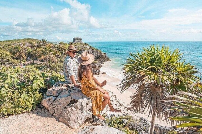 Een stel zit op een rotsachtige uitkijkpost met uitzicht op een strand, palmbomen en een helderblauwe lucht. 1
