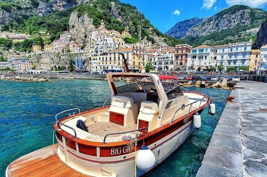 A boat named 'BIG GIO' docked at a pier with a coastal town and mountains in the background. 1