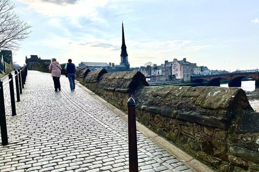 A cobblestone walkway with a stone wall, a bridge, and a tall church spire in the background. 3