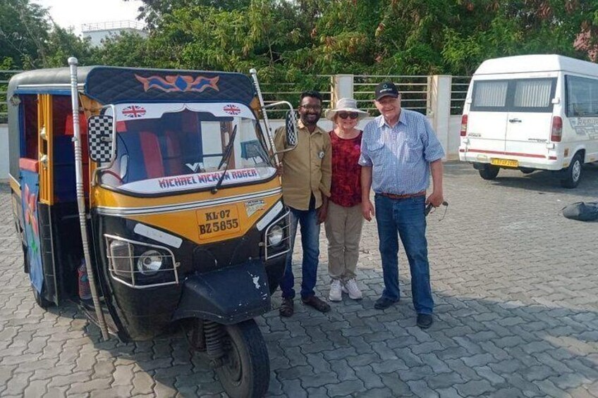 Ein gelb-blaues Auto-Rikscha mit dem Text 'MICHELE NICKLA RUGER' und dem Kennzeichen 'KL 07 BZ 5855' ist auf einer befestigten Fläche geparkt, im Hintergrund ist ein weißer Lieferwagen zu sehen. 3