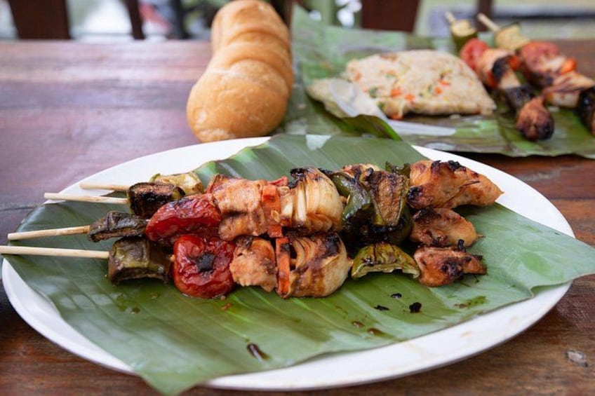 Skewered meat and vegetables on a banana leaf, with a loaf of bread in the background. 4