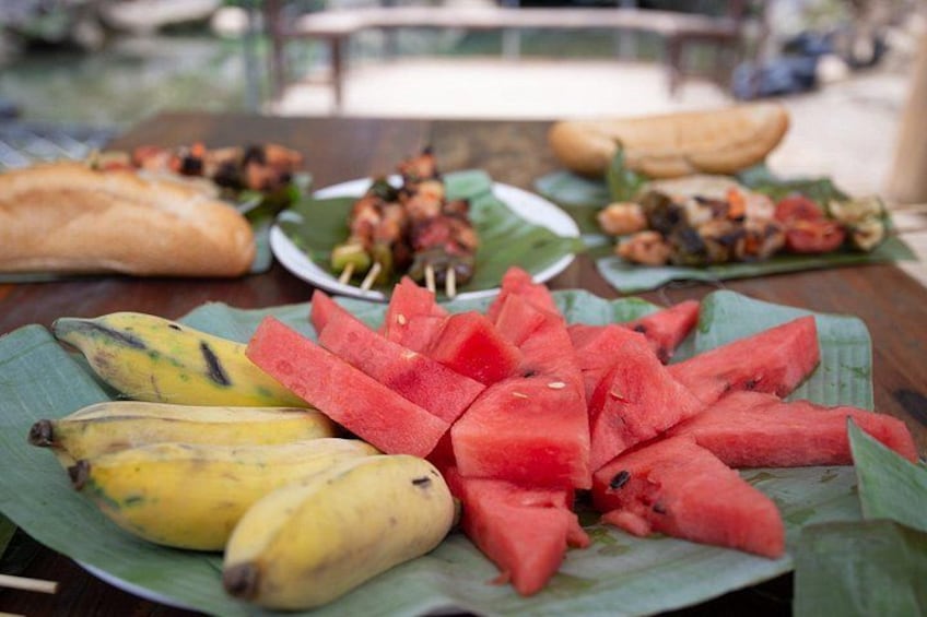 A table with sliced watermelon and bananas on a banana leaf, skewers of grilled food, and a baguette. 3