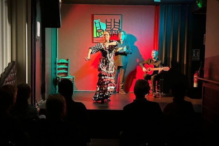 Una presentación en escenario con un cantante y dos guitarristas, una silla y un telón rojo de fondo. 2