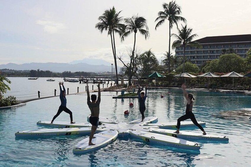 Des personnes pratiquent le yoga sur des planches de surf dans une piscine, avec des palmiers et un hôtel en arrière-plan. 4