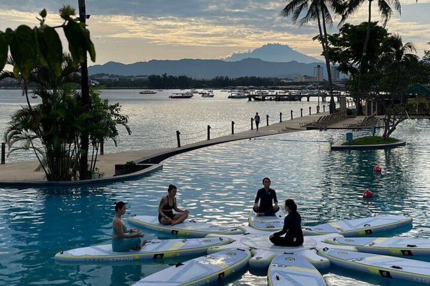 A group of people sitting on floating mats in a pool surrounded by palm trees and a dock with boats in the background. 2