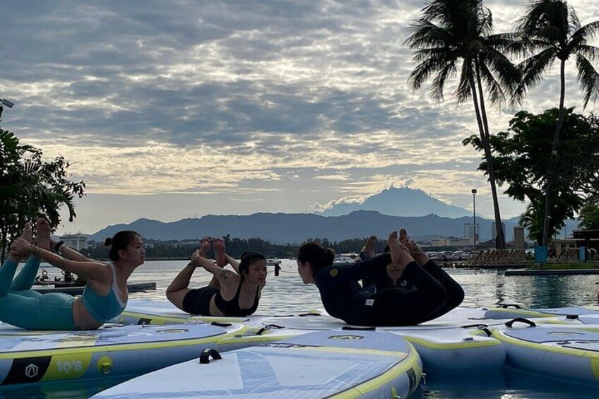 Three people doing yoga on a boat by a lake with mountains in the background. 3