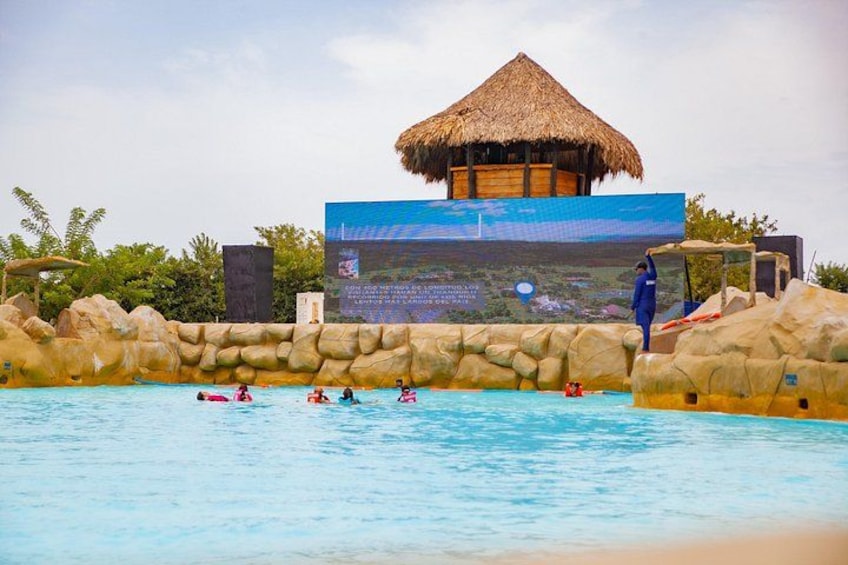 A swimming pool with people in the water, a thatched-roof structure, and a large digital display. 1