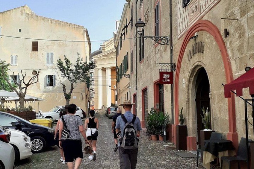 A cobblestone street with pedestrians, parked cars, and historic buildings with arches and signage. 5