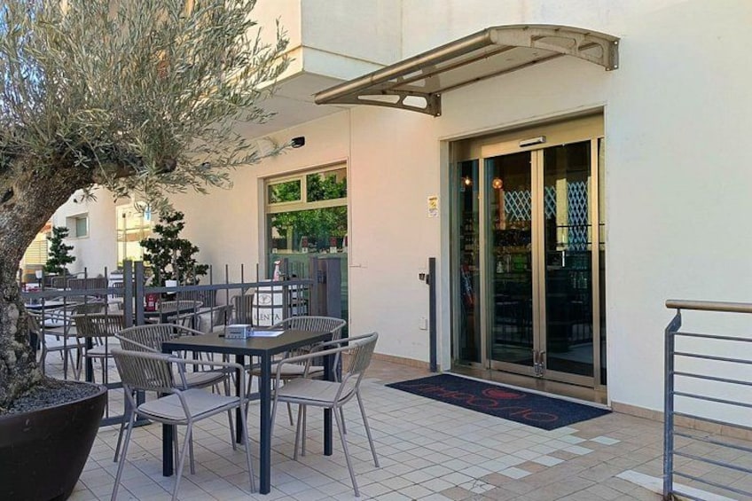 An outdoor seating area with tables and chairs, a potted tree, and a building entrance. 3