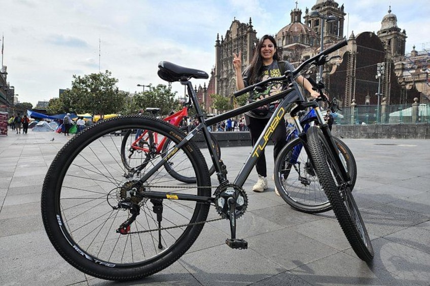 Un vélo avec un cadre noir et des détails jaunes, une personne debout à côté, et un bâtiment historique en arrière-plan. 2