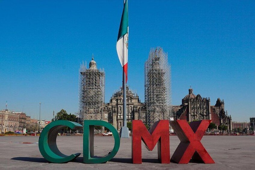Une vaste place publique, ornée des lettres « CDMX » en vert et en rouge, avec un mât portant un drapeau et un bâtiment historique entouré d’échafaudages. 4
