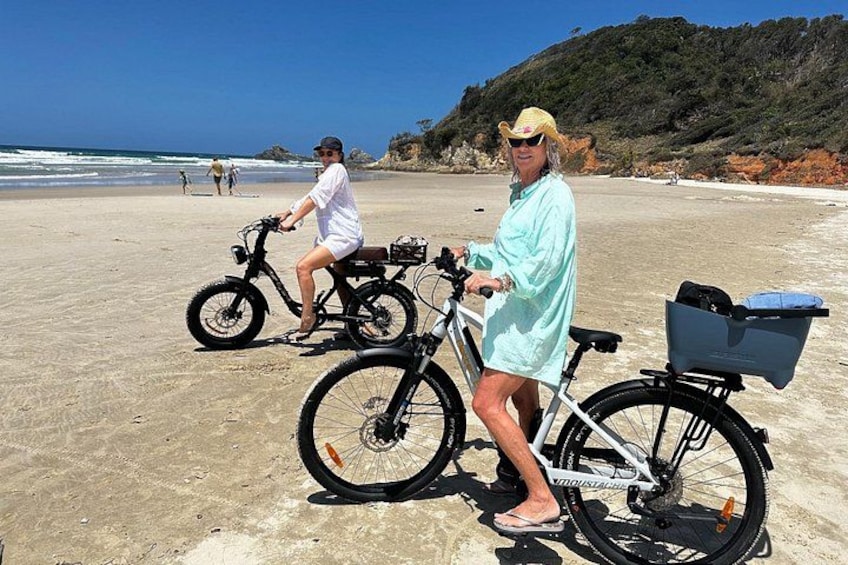 Two people on bicycles on a sandy beach with a clear sky and a hilly landscape in the background. 2