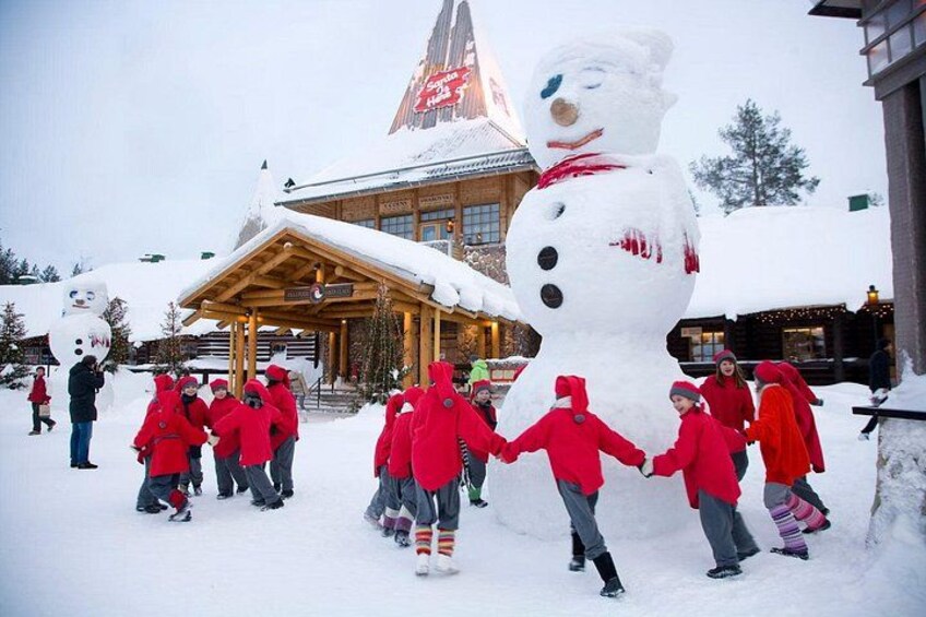 A group of people in red jackets dancing around a large snowman sculpture in a snowy outdoor setting. 4
