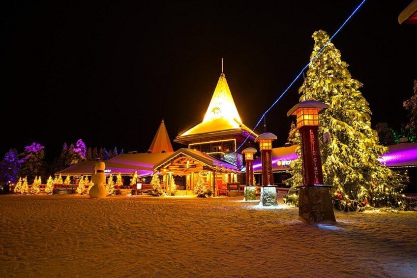 A snow-covered landscape with a lit building featuring a pointed roof and a large Christmas tree. 2