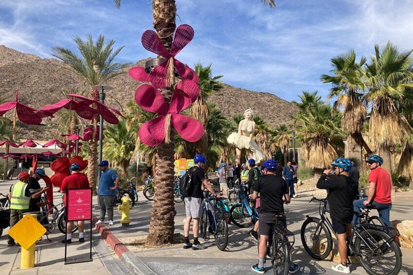Een groep fietsers verzameld rond een palmboom, versierd met een grote roze decoratie, een standbeeld en een berg op de achtergrond. 2