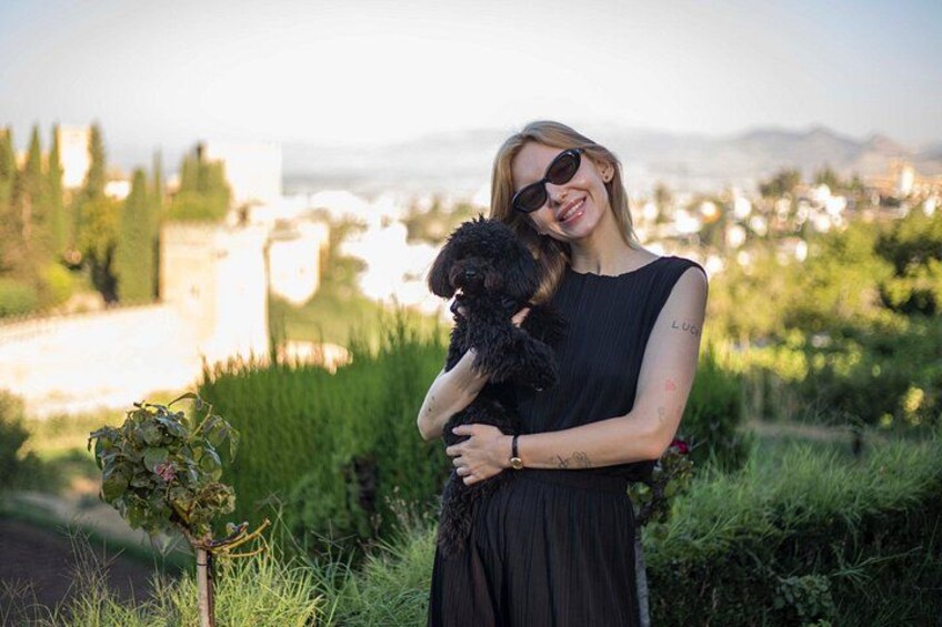 A person holding a small black dog, standing in a grassy area with a cityscape in the background. 1