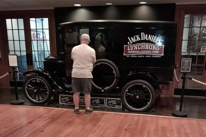 Ein altes schwarzes Auto mit dem Aufdruck 'Jack Daniel's Lynchburg Hardware & General Store', ein Mann steht davor, und ein Holzboden ist zu sehen. 1