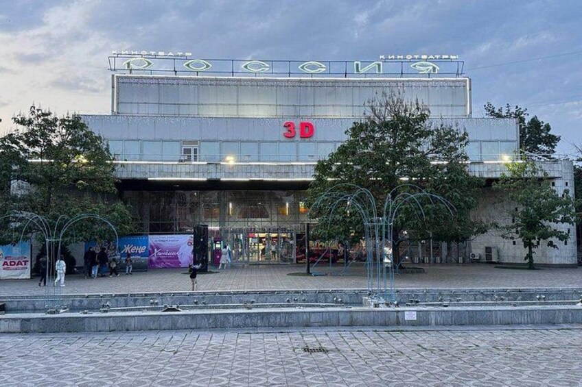 A large building with a sign reading '3D' and 'KINOASTR.' There are trees in front and people walking around. 2
