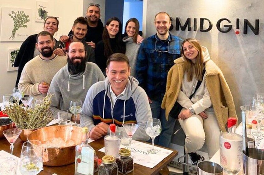 Un groupe de personnes est assis autour d’une table, devant des verres et des bouteilles de vin, dans une pièce où l’on peut voir un panneau portant l’inscription « MIDGIN » sur le mur. 4
