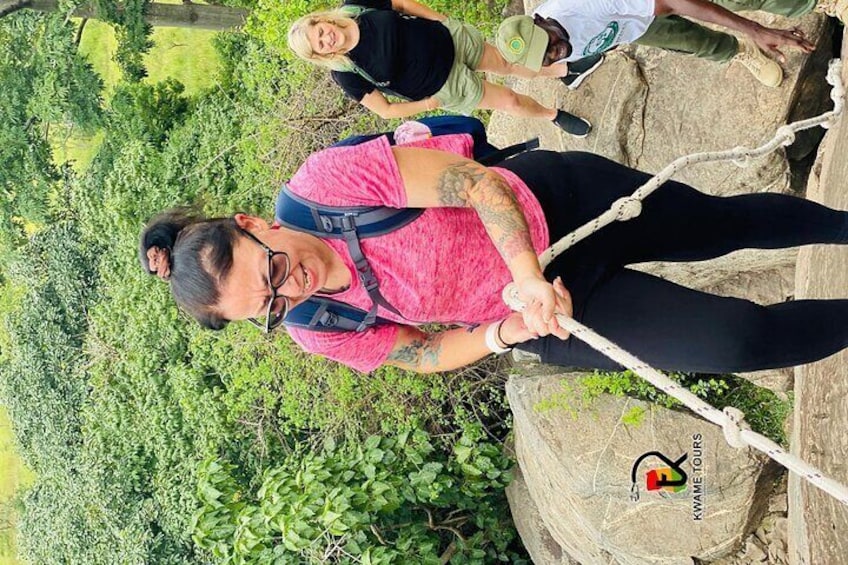 A person in a pink shirt and black pants holding a rope, standing on a rock with greenery in the background. 4