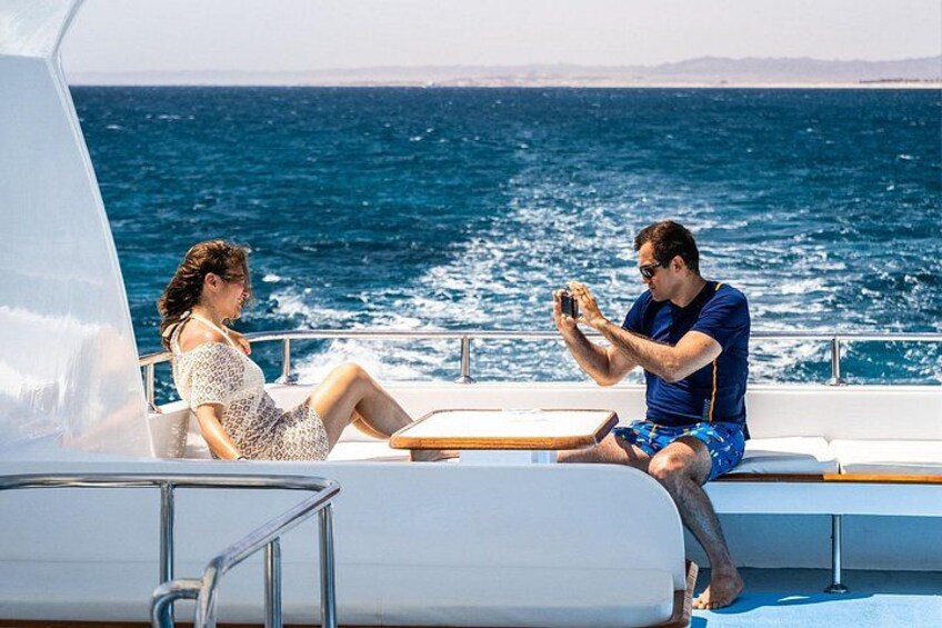Two people on a boat, one taking a photo, with the ocean and a clear sky in the background. 3