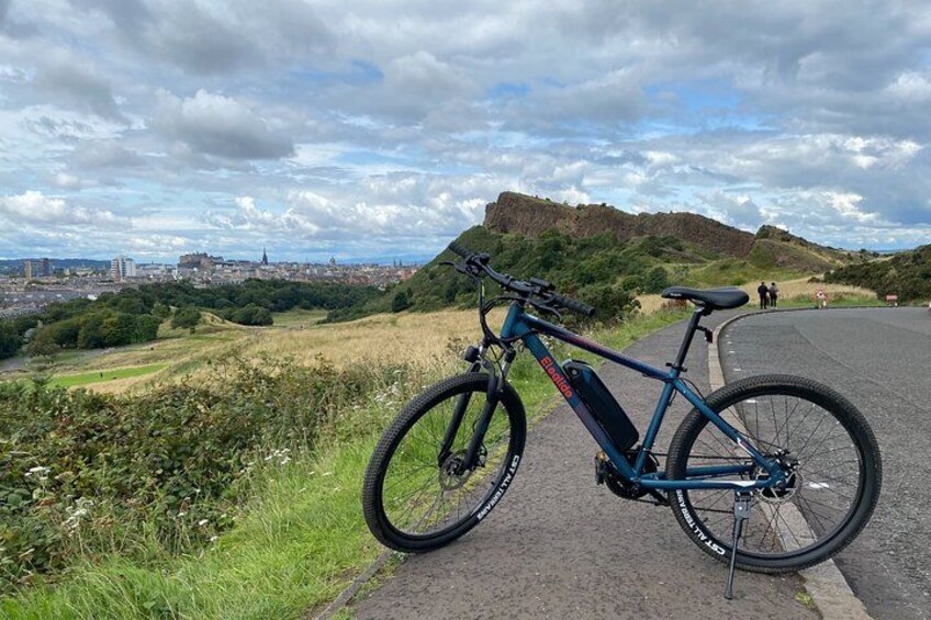 A blue electric bike parked on a paved path with a cityscape and a hill in the background. 1