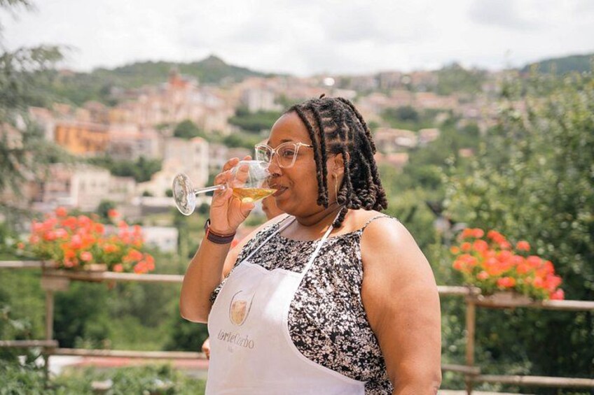 A person holding a wine glass, wearing a patterned top and an apron with a logo, standing in a garden with red flowers and a cityscape in the background. 5