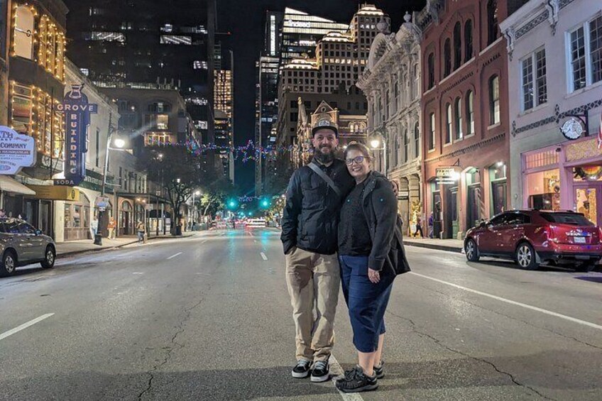 A couple standing on a city street at night with illuminated buildings and cars parked on the side. 3