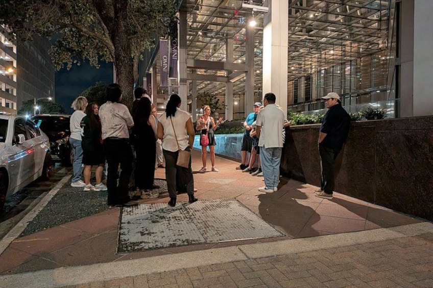 Un groupe de personnes se tient sur un trottoir, de nuit. On aperçoit un bâtiment avec une façade en verre et un arbre en arrière-plan. 5