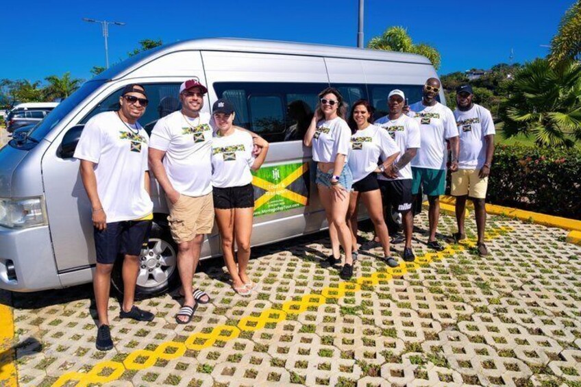 A group of people standing in front of a silver van with a green and yellow logo, on a patterned pavement with palm trees in the background. 2