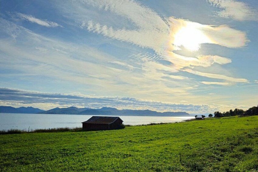A grassy field with a small wooden structure, a body of water, and mountains in the distance under a partly cloudy sky. 3