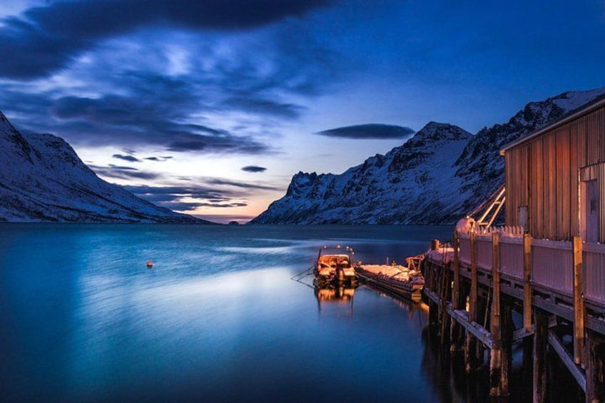 A calm fjord at dusk with a wooden dock and a building on the right, surrounded by snow-covered mountains. 1