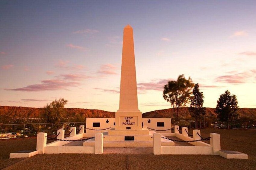 Un monumento alto y blanco con la inscripción 'LET'S FORGET' rodeado por una cerca de malla y un área pavimentada. 3