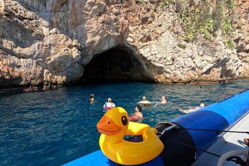 A rocky coastal area with a clear blue sea, a cave entrance, and people swimming and floating. 2