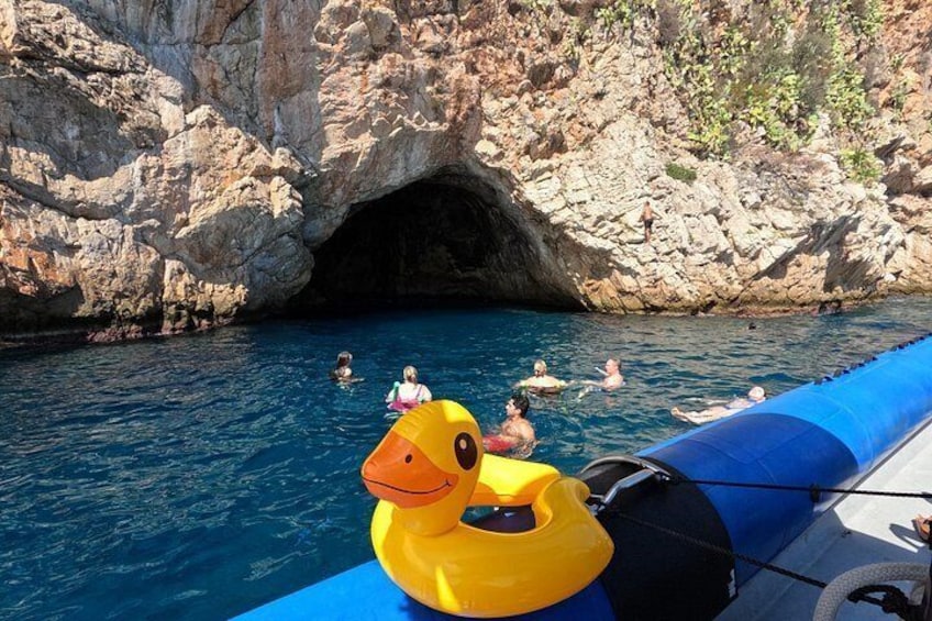 Una zona costera rocosa con mar azul cristalino, la entrada a una cueva, y gente nadando y flotando. 3