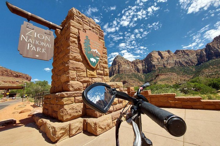 En stenkonstruktion med en skylt för Zion National Park, ett bergsmassiv i bakgrunden och en cykel i förgrunden. 1