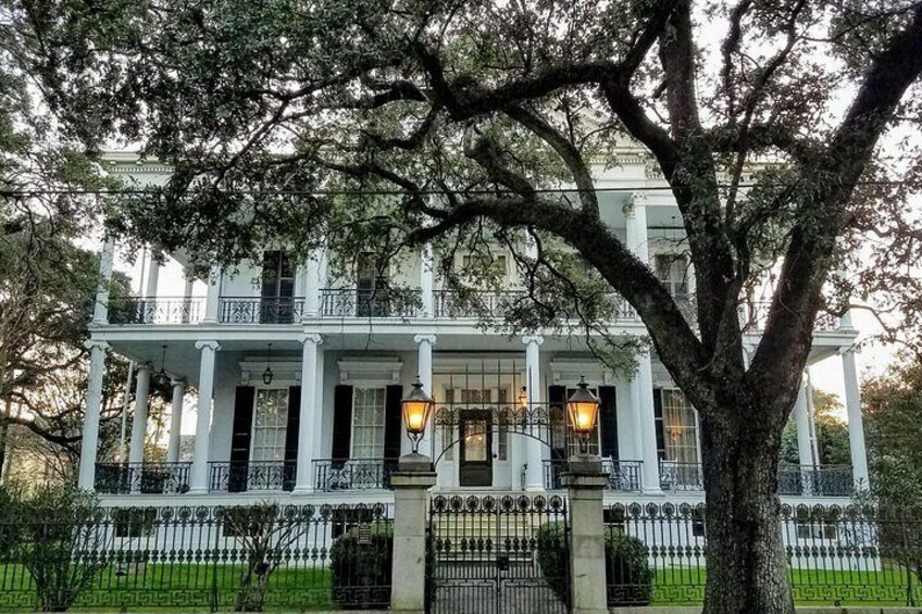 A large white building with columns and a balcony, surrounded by a fence and trees. 3