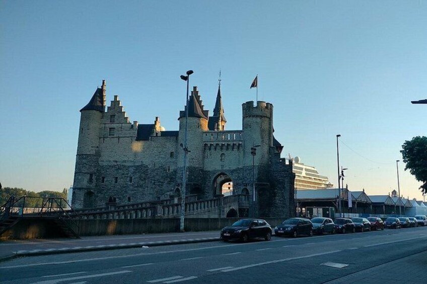 Un edificio parecido a un castillo con múltiples torres y banderas, una carretera con coches estacionados y un cielo despejado. 2