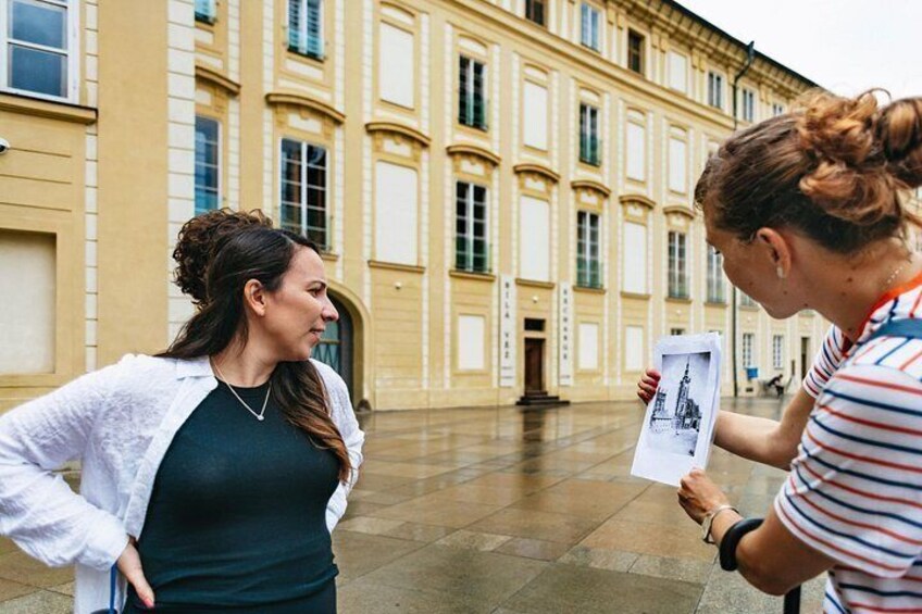 Two women standing in a courtyard with classical architecture, one holding a photo. 5