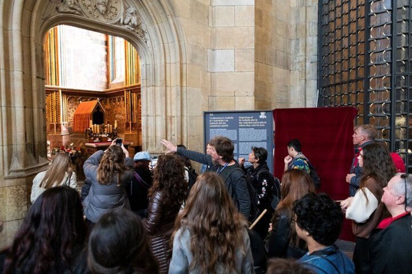 A group of people gathered inside a historic building with stone architecture, a red curtain, and a display board. 4