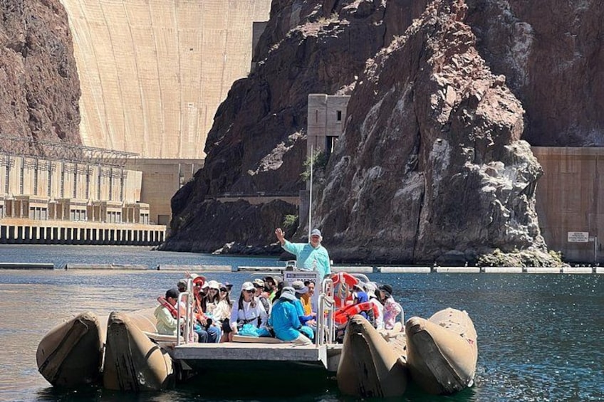 A boat tour with passengers in front of a large dam and rocky cliffs. 1