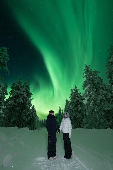 Kaksi ihmistä seisoo lumisessa maisemassa, ja taivaalla näkyy vihreä revontuli. 1