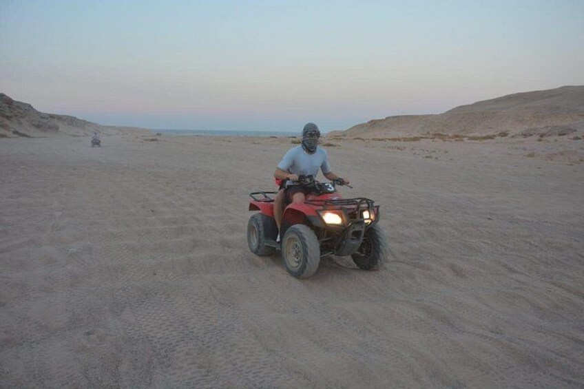 Iemand rijdt op een rode quad over een zandstrand, met duinen op de achtergrond. 5