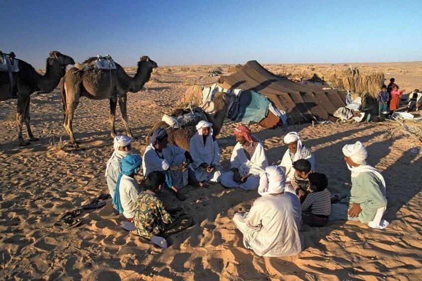 Een groep mensen zit op het zand in een woestijn, met kamelen en tenten op de achtergrond. 2