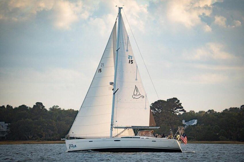A sailboat with a white hull and sails, marked with numbers and a small flag, sailing on calm water with a forested shoreline in the background. 2