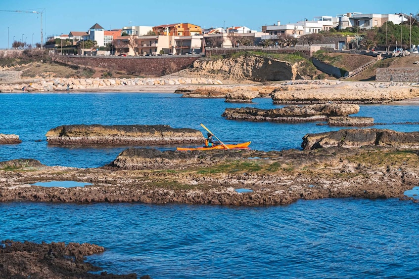 A kayaker in an orange kayak navigating rocky waters near a coastal town with buildings in the background. 2